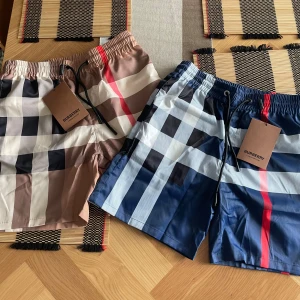 Burberry rutiga shorts - Två par Burberry shorts med klassiskt rutigt mönster, ett par i beige, vitt och brunt och ett par i blått och vitt med röda detaljer. Båda har elastisk midja och snörning. Perfekta för sommaren och ger en lyxig vibe till din outfit. 650 per st.
