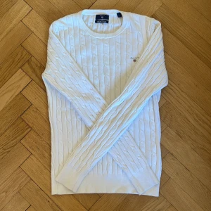 Vit kabelstickad från GANT - Säljer en klassisk vit kabelstickad tröja från GANT i stretchig bomull. Köpt för flera år sedan men knappt använd, inga fläckar eller slitage.