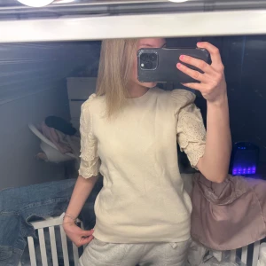 Beige stickad topp med spetsärmar - Supersöt beige stickad topp med korta puffärmar i broderad spets och volangdetaljer. Rund halsringning och ribbad mudd nertill. Perfekt för dig som gillar romantisk och trendig stil.