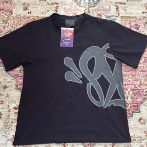 Svart t-shirt från Syna World - Svart t-shirt från Syna World med stor grå graffiti-inspirerad print som täcker både fram- och baksidan. Klassisk passform med rund hals och korta ärmar. Materialet känns mjukt och skönt, perfekt för en streetwear-look. Armarna är oversized. 