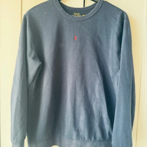 Mörkblå tröja från Polo Ralph Lauren - Klassisk mörkblå tröja från Polo Ralph Lauren i bomull med rund hals och lång ärm. Ikoniska röda broderade loggan på bröstet ger en clean och stilren look. Perfekt för dig som gillar enkel men snygg stil.