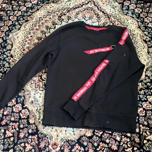 Svart Alpha Industries tröja med fack på armen. - Cool svart tröja från Alpha Industries med röda detaljer och texten 'REMOVE BEFORE FLIGHT' på banden. Tröjan har en dragkedja på ärmen och ribbade muddar. Perfekt för dig som gillar streetwear och vill sticka ut lite extra.