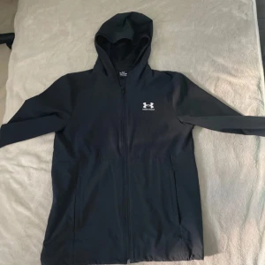  Under Armour vindjacka - Svart vindjacka från Under Armour med huva och hel dragkedja framtill. Jackan har en stilren look med logga på bröstet och praktiska fickor. Perfekt för blåsiga dagar och sportiga outfits.