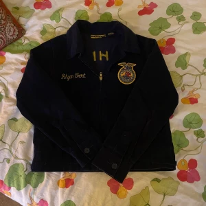 Mörkblå manchesterjacka FFA Ohio - Säljer en mörkblå manchesterjacka från FFA med broderade detaljer i gult och emblem på både fram- och baksida. Jackan har klassisk krage, dragkedja framtill och knappar vid ärmslut. Broderad text på bröstet och stor logga på ryggen!