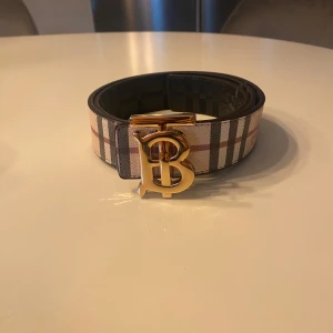 Helt nytt rutigt bälte med guldspänne.  - Snyggt bälte med klassiskt rutig design i beige, svart, vitt och rött. Bältet har ett brett band och ett stort, glänsande guldspänne. Perfekt accessoar för att lyfta din outfit med en lyxig touch.