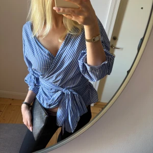 Blåvit omlottblus med knyt - Säljer en blåvit randig omlottblus med v-ringning och långa ärmar. Blusen har ett brett knytband i midjan som ger en snygg siluett och extra detalj. Perfekt för dig som gillar en fräsch och trendig look med lite volym i ärmarna.