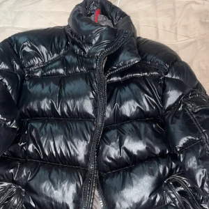 Svart pufferjacka från Moncler - Säljer en svart, glansig pufferjacka från Moncler med hög krage och dragkedja. Jackan har klassisk quiltad design och Moncler-logga på ärmen. Perfekt för kalla dagar när du vill hålla stilen och värmen. Luvan tillkommer, inga skador på den alls , använd 1 gång