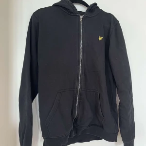 Svart hoodie från Lyle & Scott - Svart hoodie från Lyle & Scott med dragkedja och huva. Klassisk gul logga på bröstet och två stora fickor framtill. Mjuk bomullskänsla och långärmad modell, perfekt för chill dagar eller när du vill ha en enkel och stilren look.