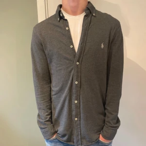 Grå långärmad skjorta Ralph Lauren - Snygg grå långärmad skjorta från Ralph Lauren i featherweight mesh. Skjortan har klassisk krage, knappar framtill och den ikoniska loggan broderad på bröstet. Perfekt för dig som gillar stilrena och bekväma plagg.