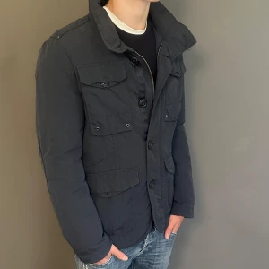 Svart pufferjacka från Belstaff - Säljer en svart pufferjacka från Belstaff med hög krage, fyra fickor framtill och snygga knappar. Jackan har dragkedja och knäppning, samt en diskret logga på ärmen. Perfekt för kyliga dagar och har en stilren, urban look.