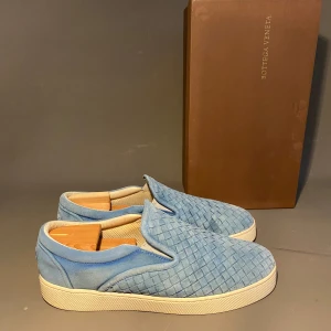 Blå slip-on sneakers från Bottega Veneta - Snygga slip-on sneakers från Bottega Veneta i ljusblå mocka med deras ikoniska flätade mönster. Skorna har vit platt sula och rund tå, vilket ger en clean och modern look. Perfekta för dig som gillar exklusiva och stilrena detaljer.