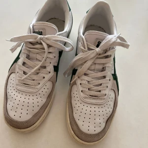 Vita Onitsuka Tiger sneakers med gröna detaljer - Snygga vita sneakers från Onitsuka Tiger med gröna detaljer och klassisk logga på sidan. Skorna har perforerad tå, snörning och beige mockadetaljer framtill. Ovandelen är i skinn och mocka, med platt sula och rund tå. Perfekta för en sportig och stilren look. St 38, använda fåtal ggr. Gott skick! 
