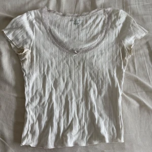 Vit ribbad topp u ringad - Supersöt vit ribbad topp med korta ärmar och spetskant runt den djupa rundade halsringningen. Små diskreta hålmönster i tyget och en liten rosett framtill. Perfekt för dig som gillar romantiska och feminina detaljer. Den är lite missfärgad i armhålorna av deodorant men inget super synligt.