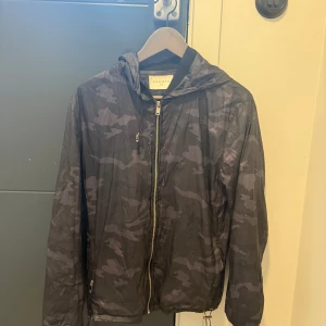 Svart camo vindjacka från Sandro - Lätt och smidig vindjacka från Sandro i svart och grått camouflagemönster. Jackan har huva, hel dragkedja framtill och dragsko nertill. Jackan är vintige och går inte att få tag på längre, köptes för 6700kr. Finns ett litet rivmärke som går att laga.