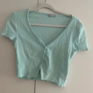 Ljusblå croppad ribbad topp/cardigan - Säljer en ljusblå croppad topp/cardigan med korta ärmar och v-ringning. Toppen är ribbad och har knappar framtill för en extra snygg detalj. Perfekt för varma dagar och enkel att matcha med jeans eller kjol