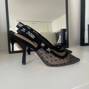 Dior J'Adior Slingback Pumps, Svarta, Storlek 38 - Dior slingback pumps i svart färg klack på 6,5cm. Dessa skor har en spetsig tå och en distinkt 'J'Adior'-detalj på bandet. Perfekt för både vardag och fest. Ligger i originalkartong samt med en sammetspåse. Använda 2 ggr. Köpta i Italien.