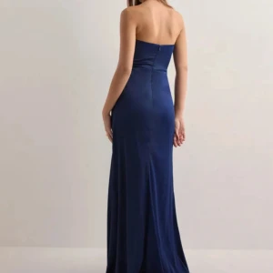 Marinblå axelbandslös långklänning - Snygg marinblå axelbandslös långklänning i glansigt material med draperad effekt vid midjan och hög slits framtill. Klänningen har en figurnära passform och stängs med dragkedja i ryggen. Perfekt för dig som vill sticka ut med en elegant look. Köpt för ordinarieprid 800kr