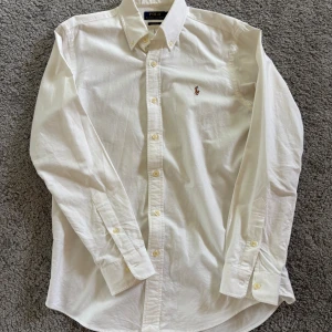 Vit skjorta från Polo Ralph Lauren - Klassisk vit skjorta från Polo Ralph Lauren i bomull med broderad logga på bröstet. Skjortan har lång ärm, knappar hela vägen och button down-krage. Custom fit-modell som sitter snyggt och passar till många olika stilar. Den enda defekten är gulnad runt armhålor och hals! 