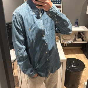 Jeansskjorta från Ralph Lauren - Säljer en snygg jeansskjorta från Ralph Lauren i slim fit. Skjortan är blå med klassisk krage, knappar hela vägen och ikoniska broderade loggan på bröstet. Perfekt för dig som gillar en stilren och avslappnad look.