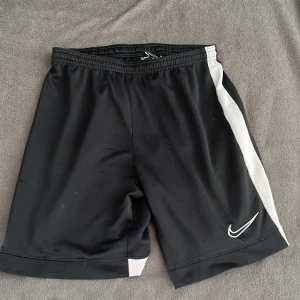 Svarta Nike Dri-Fit shorts L - Svarta sportiga Nike Dri-Fit shorts med vita sidor och logga framtill. De har elastisk midja med snörning för perfekt passform. Tillverkade i ett lätt och snabbtorkande syntetmaterial, perfekta för träning eller fotboll. Klassisk loose fit och stilren design.