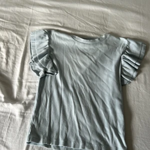 Ljusblå volangärmad topp Gina Tricot - Supersöt ljusblå topp från Gina Tricot med ribbad struktur och volangdetaljer på ärmarna. T-shirten har rund halsringning och korta ärmar med extra volym. Perfekt för dig som gillar en feminin och trendig stil.