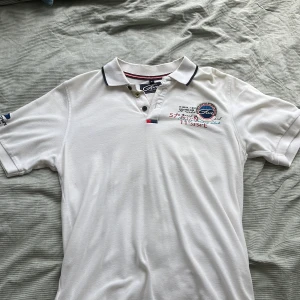 Vit pikétröja från Five Seasons M - Vit pikétröja från Five Seasons med broderad logga och text på bröstet. Klassisk krage med knappar och snygga detaljer i rött och marinblått på insidan av kragen. Perfekt för en sportig och stilren look.