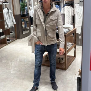 Beige vindjacka med fickor och dragkedja - Helt ny beige vindjacka med prislappen kvar. Jackan har en klassisk krage och är perfekt för dig som vill ha en stilren och funktionell look. Tillverkad i lätt syntetmaterial som passar bra till jeans eller chinos.