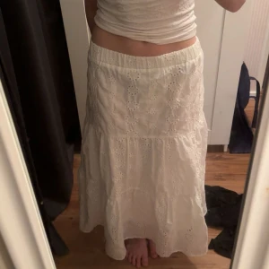 Vit långkjol med broderade detaljer - Superfin vit långkjol med broderade blommor och hålmönster. Kjolen har resår i midjan och faller snyggt i flera lager. Perfekt för sommarens alla tillfällen och ger en romantisk vibe.