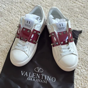 Valentino Rockstud vita sneakers - Snygga vita Valentino Rockstud sneakers med breda röda band över vristen dekorerade med ikoniska silverfärgade nitar. Skorna har rund tå, vita snören och är tillverkade i skinn.