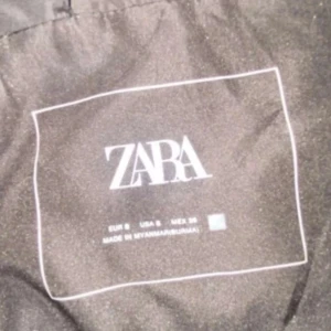 Svart dunväst med huva från Zara - Säljer en svart dunväst från Zara med snygg quiltad design och stor huva. Västen har dragkedja framtill och är perfekt för lager-på-lager-stil. Passformen är normal och materialet är mjukt och lätt, vilket gör den enkel att bära över en hoodie eller tröja.