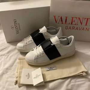 Valentino Garavani Open sneakers vit/svart - Snygga Valentino Garavani Open sneakers i vitt skinn med bred svart läderdetalj över vristen. Klassisk rund tå, vita snören och platt sula med ikoniska nitar baktill. Perfekta för dig som vill ha en clean men lyxig look.Allt og finns kvar