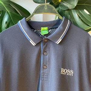 Hugo boss pikétröja  - Snygg mörkblå pikétröja från Hugo Boss med vit logga broderad på bröstet. Klassisk krage med vita och blå ränder, tre knappar framtill och korta ärmar. Perfekt för en clean och stilren look. Tillverkad i mjuk bomull för skön känsla.