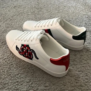 Gucci Ace vita sneakers med orm - Gucci Ace sneakers i vitt skinn med klassiska gröna och röda ränder på sidan och en broderad röd orm. Baksidan har metallicröd och grön detalj med Gucci-logga. Snygg och ikonisk modell med snörning och platt sula.