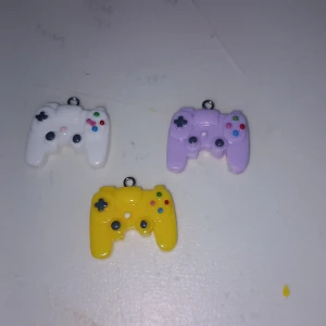 Färgglada halsbandshängen i form av spelkontroller - Tre coola halsbandshängen formade som spelkontroller i färgerna vit, lila och gul. Perfekta för dig som älskar gaming och vill ha något unikt och lekfullt i din stil. Hängena har små färgglada knappar och passar perfekt på en kedja eller nyckelring.