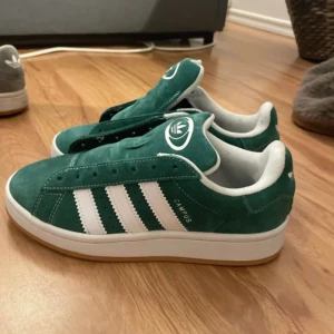 Adidas Campus gröna sneakers mocka - Gröna Adidas Campus sneakers i mocka med klassiska vita ränder på sidorna och vit logga på häl och plös. Skorna har vit insida och en snygg gummisula i ljusbrunt. Perfekta för dig som gillar retrostil och streetwear.