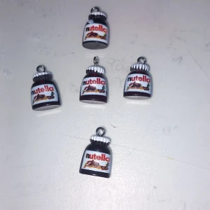 Nutella-berlocker till smycken - Fem små berlocker formade som Nutella-burkar, perfekta att använda till halsband, armband eller örhängen. De är bruna med vita lock och har detaljerad etikett med Nutella-loggan. Supergulliga och unika accessoarer för dig som älskar Nutella!