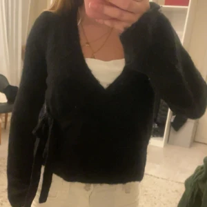 Svart omlottkofta från Gina Tricot - Mysig svart omlottkofta från Gina Tricot i storlek S. Koftan har knytband i sidan, v-ringning och långa ärmar. Perfekt att slänga på sig över ett linne eller t-shirt. Materialet är stickat och känns riktigt skönt mot huden.