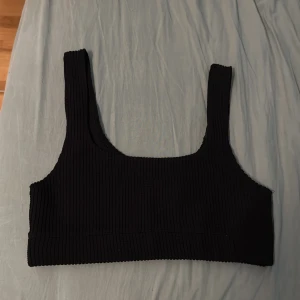 Svart ribbad linnetopp från BikBok - En svart ribbad linnetopp med breda axelband och fyrkantig urringning. Toppen är croppad och har en tight passform som framhäver siluetten. Perfekt att styla med höga jeans eller kjol för en clean look.