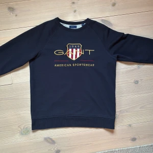 Mörkblå sweatshirt från GANT - Snygg mörkblå sweatshirt från GANT med broderad logga och sköld framtill i guld, rött och vitt. Klassisk rund halsringning och långa ärmar. Mjuk insida och ribbade muddar vid ärmslut och nederkant. Perfekt för en sportig och stilren look. Priset kan diskuteras vid snabb affär. 