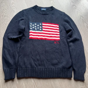 Mörkblå stickad tröja Polo Ralph Lauren - Mörkblå stickad tröja från Polo Ralph Lauren med stor amerikansk flagga framtill och RL-broderi. Klassisk rund halsringning och ribbade muddar. Perfekt statement-piece för dig som gillar preppy stil och vill sticka ut. Längd 63 cm och Bredd 43,5. Storlek xs
