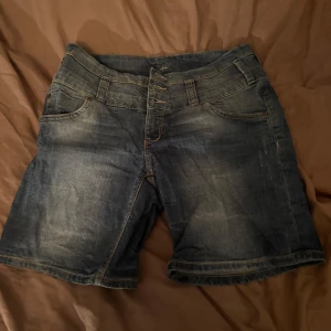Blå jeansshorts med knappar - Snygga blå jeansshorts med flera knappar framtill och klassiska fickor både fram och bak. Shortsen har en lite längre modell och coola slitningar för en avslappnad look. Perfekta för sommardagar och passar till allt i garderoben.