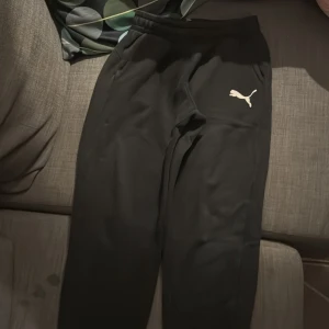 Svarta mjukisbyxor från Puma - Svarta mjukisbyxor från Puma med klassisk vit logga på vänster lår. Byxorna har resår i midjan och sidofickor, perfekta för chill eller träning. Tillverkade i mjukt och bekvämt material som känns skönt mot huden.