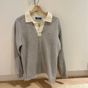 Grå pikétröja från Adidas Originals - Snygg grå pikétröja från Adidas Originals med vit krage och knappslå. Tröjan har broderad Adidas-logga på bröstet och är långärmad. Perfekt för en sportig och avslappnad stil. Tillverkad i mjuk bomull för extra komfort.*mjukis material* 