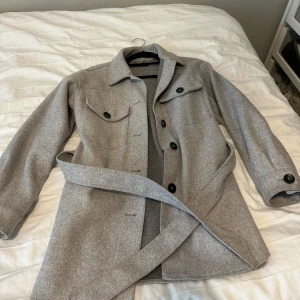 Grå overshirt med bälte från Zara - Snygg grå overshirt från Zara i ullmix med bälte i midjan och svarta knappar. Jackan har två bröstfickor med lock och klassisk krage. Perfekt för lager-på-lager och chill höststil.