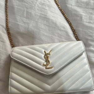 Vit quiltad axelväska från Saint Laurent - Säljer en snygg vit axelväska från Saint Laurent med guldiga detaljer och kedjerem. Väskan är quiltad i ett stilrent mönster och har YSL-logga framtill. Invändigt finns flera fack och en dragkedja för extra förvaring. Köpt för 3000 kr 