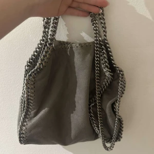 Grå axelväska Stella McCartney - Säljer en snygg grå axelväska från Stella McCartney med kedjedetaljer runt kanterna och som axelrem. Väskan är i mjuk mocka och har en cool, modern look. Perfekt storlek för det viktigaste och ger en edgy vibe till din outfit. Det är mellan storleken alltså den som är lite större än den minsta.