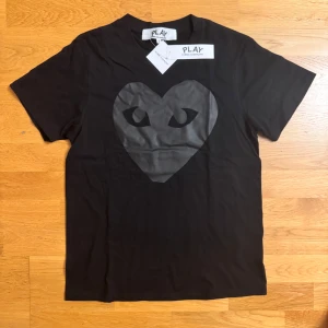 Svart t-shirt Comme des Garçons Play - Svart t-shirt från Comme des Garçons Play med det ikoniska hjärta-trycket i mörkgrått på bröstet. Klassisk passform, rund hals och korta ärmar. Tillverkad i mjuk bomull, perfekt för en clean och stilren look.
