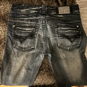 Blå bootcut jeans med nitar  - Säljer ett par blå bootcut jeans från Bianca Denim med slitna detaljer och coolt mönster på bakfickorna. Jeansen har en liten ficka framtill med silvriga nitar som ger extra edge. Klassisk femficksmodell och snygg tvättad look. Mått: Innerbenslängden 84-85cm,                      utebenslängd 104-105cm