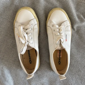 Vita Superga sneakers i skinn - Klassiska vita Superga sneakers i skinn med snörning och platt sula. Skorna har rund tå och diskret Superga-logga på sidan. Yttersulan är i gummi och har en beige ton. Perfekta för en clean och stilren look.