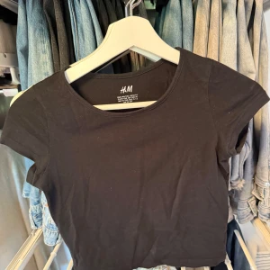 Svart croppad topp från H&M - Enkel och stilren svart croppad topp från H&M med rund halsringning och korta ärmar. Perfekt att matcha med jeans eller kjol för en clean look. 